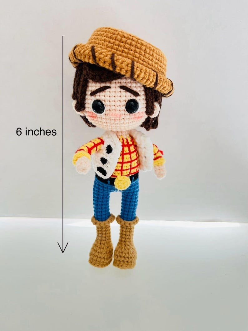 May include: A crocheted toy figure of a cowboy, standing 6 inches tall. The figure is wearing a brown hat, a red and yellow plaid shirt, blue jeans, and brown boots. The figure has brown hair and a friendly face.