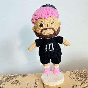 May include: A handmade crochet doll featuring a figure with bright pink textured hair, a brown beard, and a winking expression. The doll wears a black short-sleeved shirt with the number 10 embroidered in white, black shorts, and pink shoes.