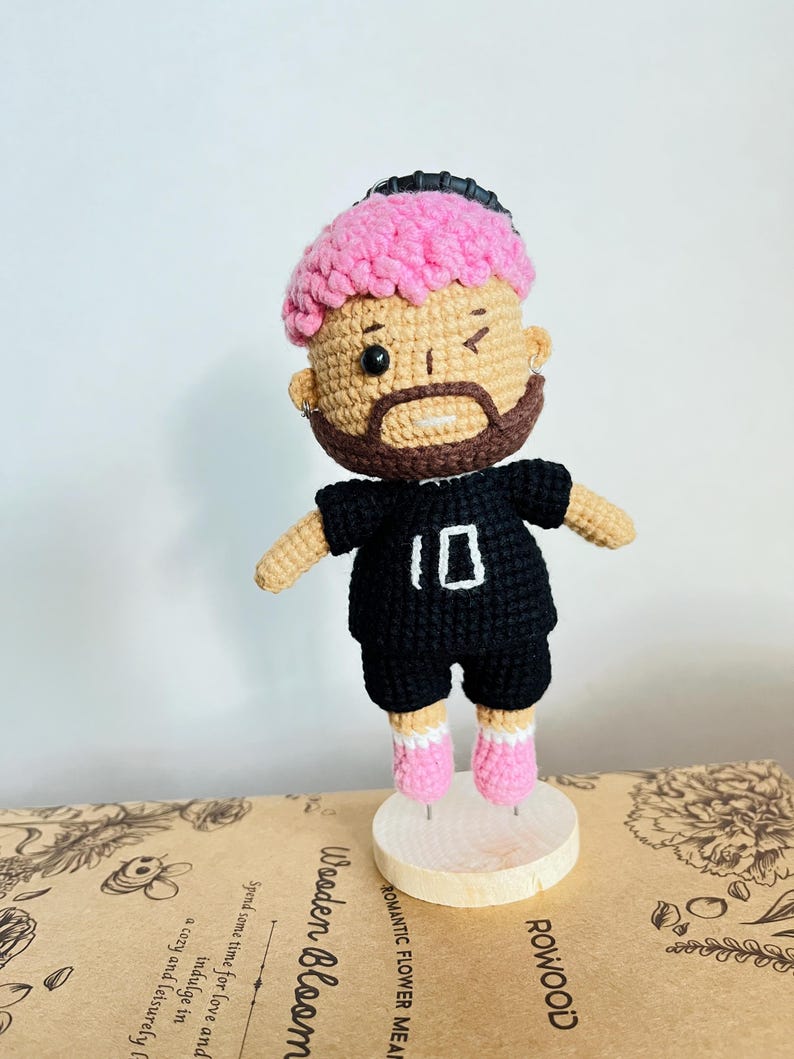 May include: A handmade crochet doll featuring a figure with bright pink textured hair, a brown beard, and a winking expression. The doll wears a black short-sleeved shirt with the number 10 embroidered in white, black shorts, and pink shoes.