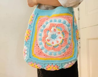 May include: A crocheted tote bag with a colorful floral design in shades of pink, blue, yellow, and white. The bag has a long strap and is made with a granny square pattern.