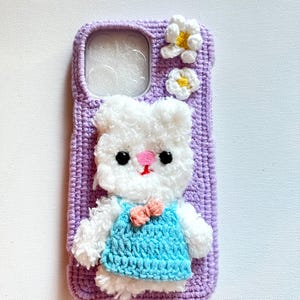 Puede incluir: Una funda de tel&eacute;fono de crochet morado con un conejito de crochet blanco que lleva un vestido de crochet azul y un lazo rosa. La funda tiene dos flores de crochet blancas con centros amarillos.