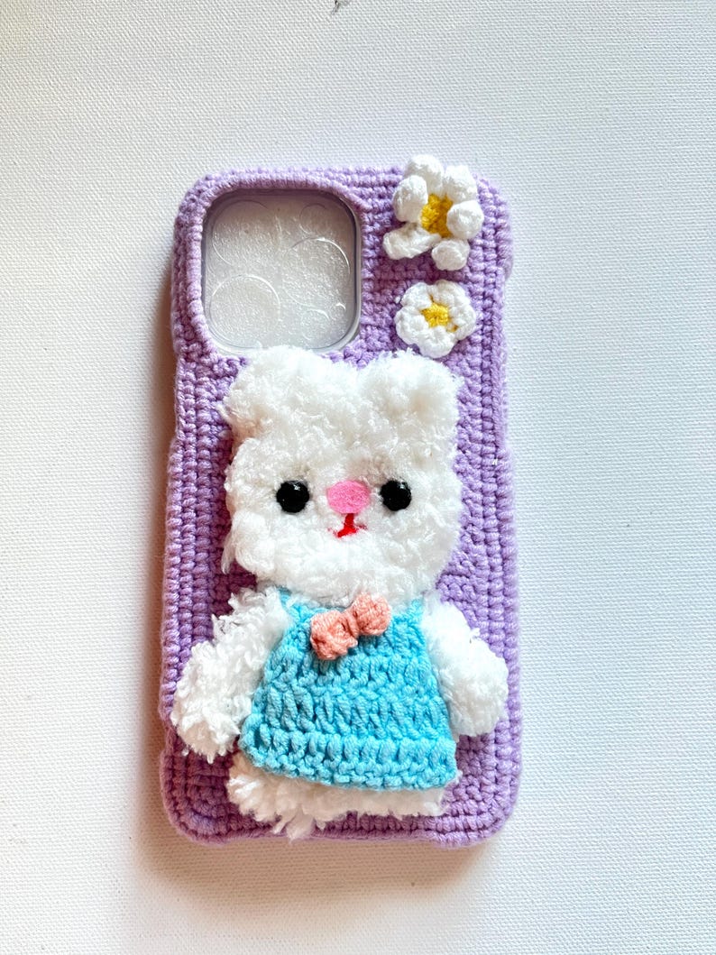 Puede incluir: Una funda de tel&eacute;fono de crochet morado con un conejito de crochet blanco que lleva un vestido de crochet azul y un lazo rosa. La funda tiene dos flores de crochet blancas con centros amarillos.