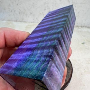 May include: A rectangular block of stabilized wood featuring a vibrant, iridescent finish with alternating horizontal bands of deep purple, royal blue, and emerald green. The surface displays a distinct wavy grain pattern throughout the material.