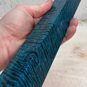 May include: A rectangular block of wood with a vibrant blue and black striped pattern. The wood grain creates a textured appearance. The block is held in a hand, showcasing the color and texture of the wood.