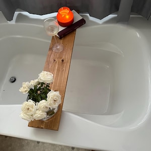 May include: A wooden bath tray with a glass of wine, a lit candle, and a bouquet of white roses. The tray is placed in a white bathtub.