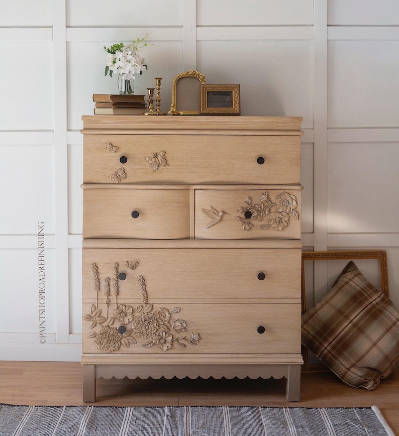 May include: A light-colored wooden dresser with five drawers and decorative floral and butterfly carvings. The dresser has black knobs and a scalloped base. A pillow and picture frames are next to the dresser.