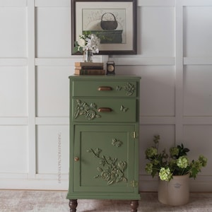 Puede incluir: Un mueble verde salvia con tres cajones y una puerta, adornado con motivos florales y mariposas. El mueble tiene herrajes color bronce y descansa sobre patas de madera oscura. Una imagen enmarcada y flores encima.