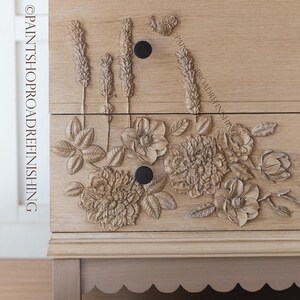 May include: A light-colored wooden dresser with carved floral and wheat stalk designs. The dresser has two drawers with black knobs and a scalloped base. The text "@PAINTSHOPROADREFINISHING" is visible on the side.