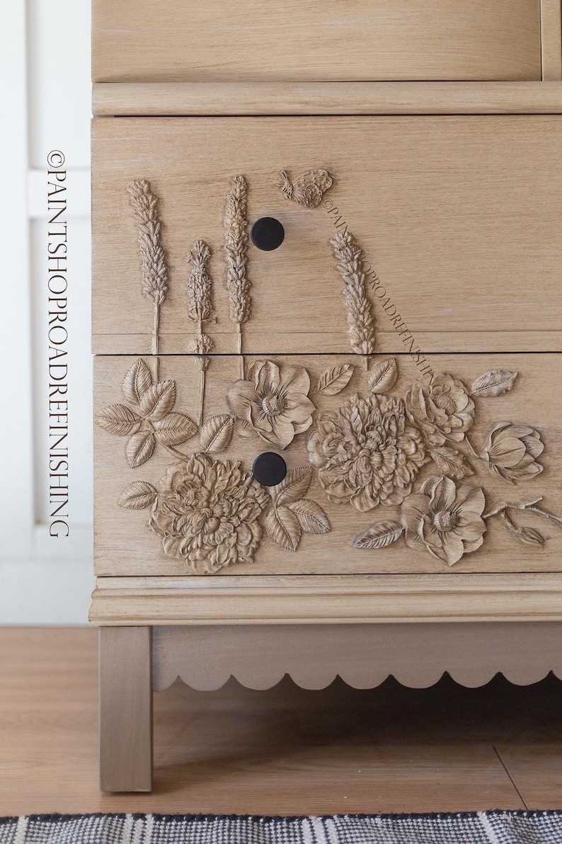 May include: A light-colored wooden dresser with carved floral and wheat stalk designs. The dresser has two drawers with black knobs and a scalloped base. The text "@PAINTSHOPROADREFINISHING" is visible on the side.