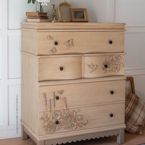 May include: A light-colored wooden dresser with four drawers, adorned with butterfly and floral carvings. The dresser has black knobs and sits on short, light-colored legs. Decorative items are on top.