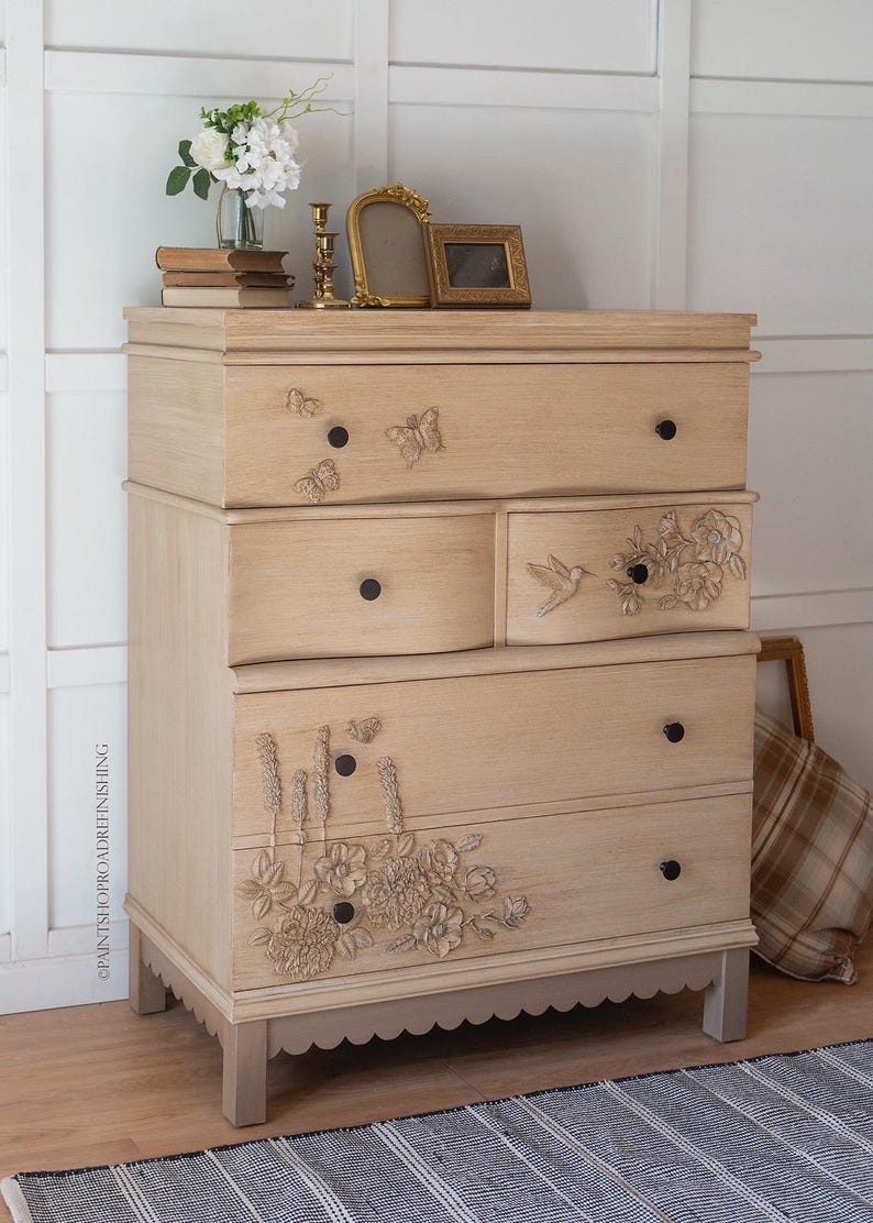 May include: A light-colored wooden dresser with four drawers, adorned with butterfly and floral carvings. The dresser has black knobs and sits on short, light-colored legs. Decorative items are on top.