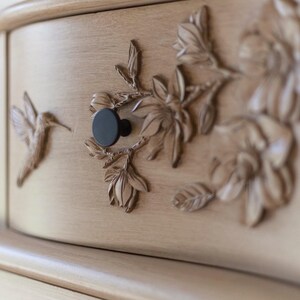 May include: Close-up of a light-colored wooden cabinet drawer with carved floral and hummingbird details. The drawer features a black knob and intricate relief carvings of flowers and a hummingbird. The cabinet has a curved front.