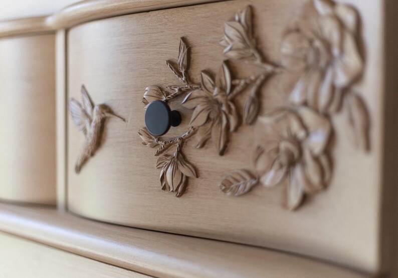 May include: Close-up of a light-colored wooden cabinet drawer with carved floral and hummingbird details. The drawer features a black knob and intricate relief carvings of flowers and a hummingbird. The cabinet has a curved front.