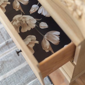 May include: A partially opened wooden drawer reveals a floral pattern on a dark background. The drawer is part of a light-colored wooden dresser. The floral design features large, cream-colored flowers.