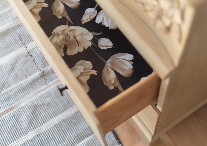 May include: A partially opened wooden drawer reveals a floral pattern on a dark background. The drawer is part of a light-colored wooden dresser. The floral design features large, cream-colored flowers.