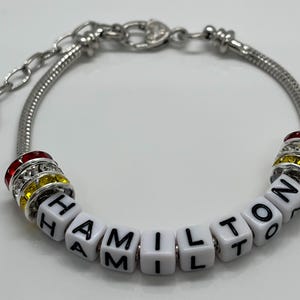 May include: A silver charm bracelet with the name "HAMILTON" spelled out in white beads with black lettering. The bracelet has red and yellow crystal beads.