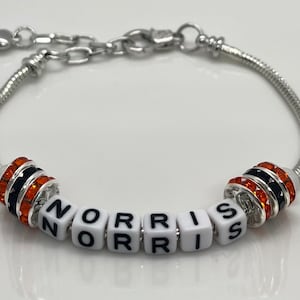Peut inclure: Un bracelet à breloques argenté avec des perles de cristal noires et orange et des perles de lettres blanches épellant le nom "NORRIS".