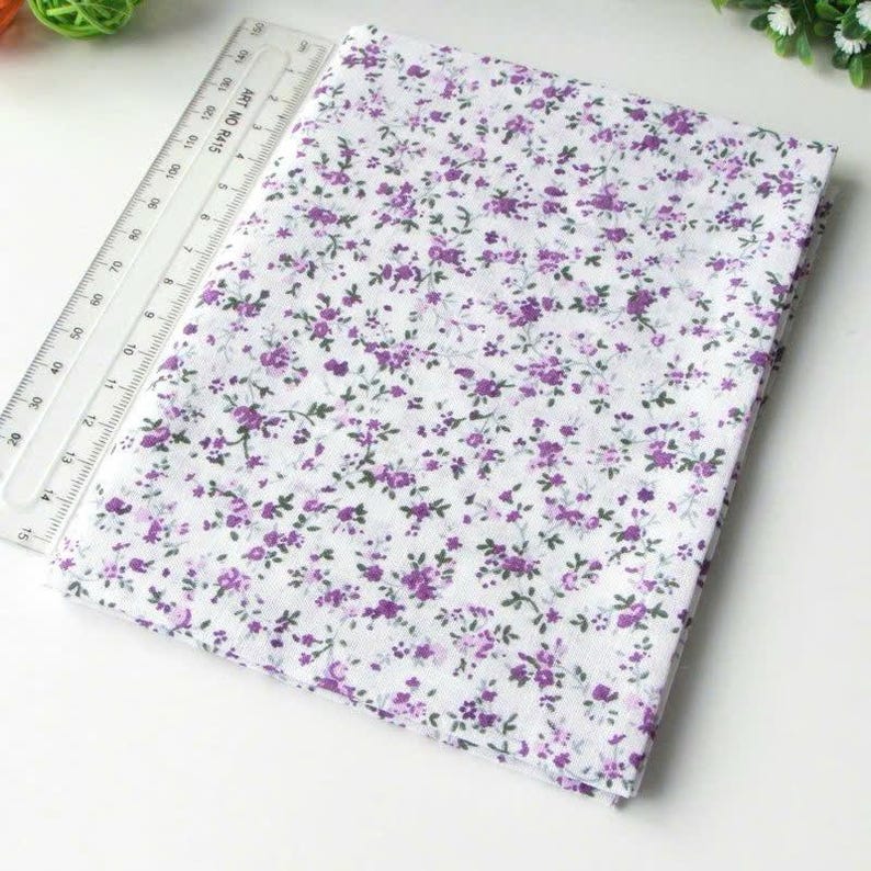 May include: A folded piece of white fabric featuring a small purple floral pattern with green stems. A clear plastic ruler is placed alongside the fabric, showing a measurement scale that extends up to 15 inches in length.