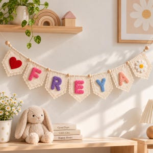 May include: A decorative fabric banner featuring a series of pennants with lace trim, displaying the name FREYA in colorful tufted letters. The banner is accented with a red heart on the left, a white daisy on the right, and wooden beads.