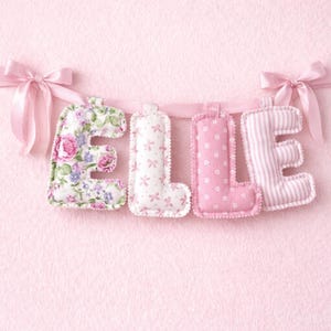 Op de afbeelding: Een roze banner met de tekst "ELLE" met stoffen letters. De "E" is bloemig, de "L"'s zijn versierd met strikken en sterren, en de laatste "E" heeft roze en witte strepen. De banner is geregen met een roze lint en strikken.