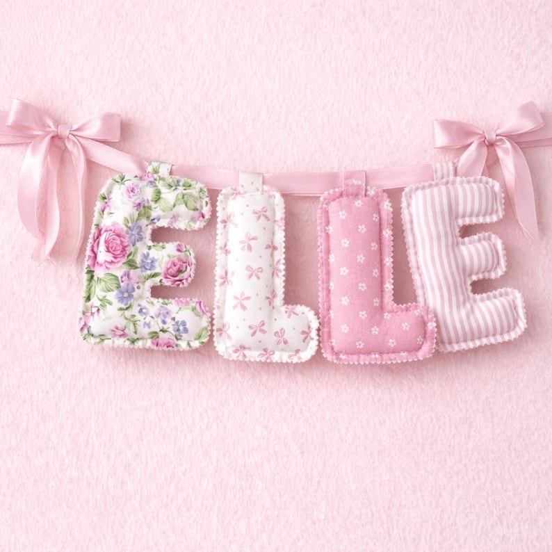 May include: A fabric banner spelling the name ELLIE with four stuffed, plush letters. The E features a pink and purple floral print, the Ls have a white background with pink bows and small pink flowers, and the E is pink with white stripes.
