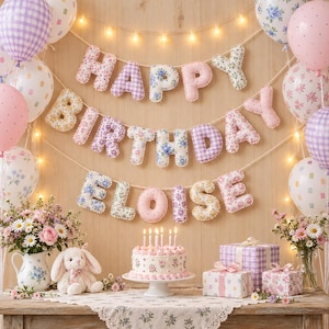 May include: A fabric banner spelling HAPPY BIRTHDAY ELOISE with stuffed, patterned letters in pastel colors. The display includes balloons, a floral cake with six lit candles, a plush rabbit, and two wrapped gifts on a wooden table.