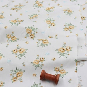 May include: A white fabric featuring a repeating pattern of yellow roses with green leaves and small light blue bows. A single wooden spool is placed on the surface of the fabric, which is folded to show the floral print clearly.