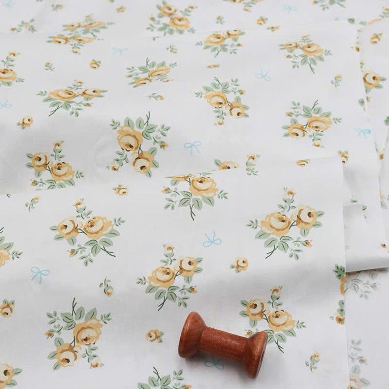 May include: A white fabric featuring a repeating pattern of yellow roses with green leaves and small light blue bows. A single wooden spool is placed on the surface of the fabric, which is folded to show the floral print clearly.