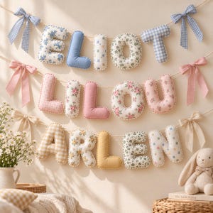 May include: A decorative fabric banner featuring three rows of stuffed, plush letters spelling ELIOT, LILOU, and ARLEN. The letters are crafted from various patterned and solid-colored fabrics, accented with bows on the ends of each string.