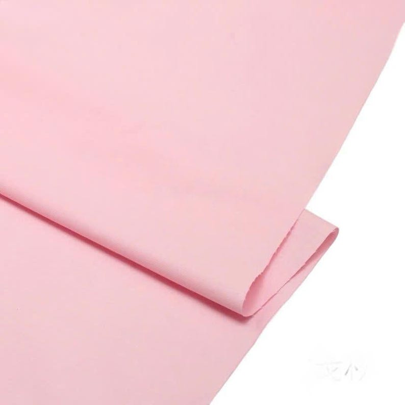 May include: A solid light pink fabric with a smooth, matte texture. The material is folded over itself, creating a soft diagonal crease across the surface. The color is consistent throughout the entire visible piece of textile.