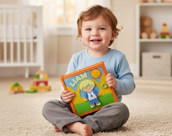 Può includere: Un bambino piccolo seduto su un tappeto tiene in mano un libro colorato. Il libro presenta il nome "LIAM" su sfondo blu, un sole e un personaggio dei cartoni animati. Il libro ha bordi verdi e arancioni. Il bambino indossa una maglietta blu a maniche lunghe e pantaloni grigi.