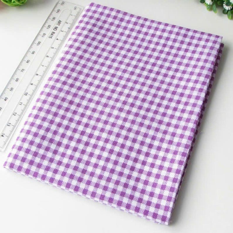 May include: A folded piece of purple and white gingham fabric featuring a small checkered pattern. A clear plastic ruler is placed alongside the fabric, showing a length measurement of approximately 15 centimeters along the edge.