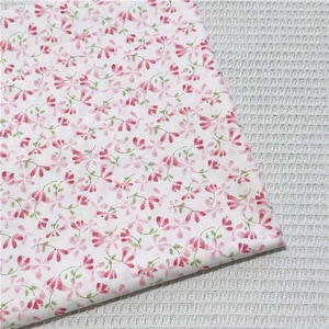 May include: A white fabric featuring a repeating pattern of small pink flowers with delicate green stems and leaves. The textile has a smooth surface texture and is displayed against a light gray waffle-weave fabric background.