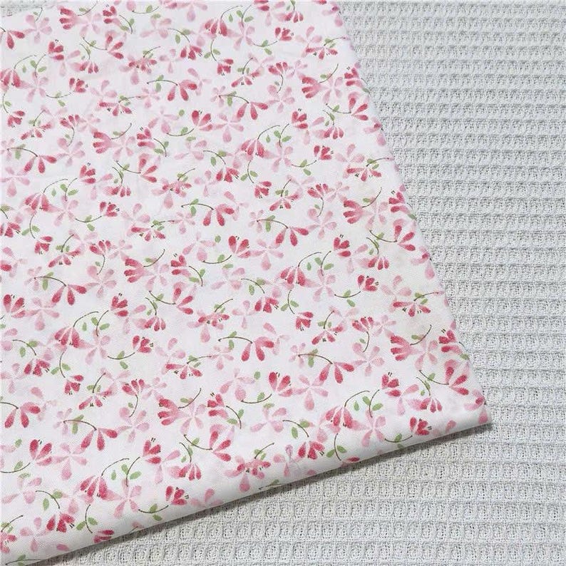 May include: A white fabric featuring a repeating pattern of small pink flowers with delicate green stems and leaves. The textile has a smooth surface texture and is displayed against a light gray waffle-weave fabric background.