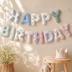 May include: A fabric banner spelling HAPPY BIRTHDAY with stuffed, plush letters in various floral, solid blue, and solid green patterns, strung on a white cord. A beige felt crown with the number 2 sits on a wooden stool nearby.