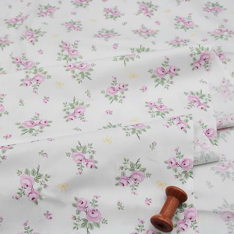 May include: A white fabric featuring a repeating pattern of small pink roses with green leaves and tiny yellow bows. A single wooden spool is placed on the surface of the folded textile, which displays soft drapes and folds across the frame.