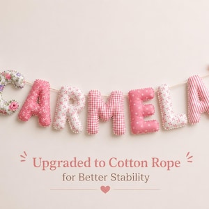 May include: A fabric letter garland spelling the name CARMELA in various shades of pink and white patterns, including floral, gingham, and polka dots. The letters are strung on a rope with pink ribbon bows at each end. Text reads: Upgraded to Cotton Rope for Better Stability.