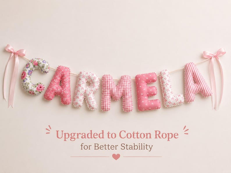 May include: A fabric letter garland spelling the name CARMELA in various shades of pink and white patterns, including floral, gingham, and polka dots. The letters are strung on a rope with pink ribbon bows at each end. Text reads: Upgraded to Cotton Rope for Better Stability.
