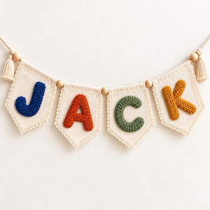 May include: A decorative banner featuring four off-white fabric pennants suspended on a cord with wooden beads and tassels. Each pennant displays a large, textured, knitted letter in blue, orange, green, and yellow, spelling out the name JACK.