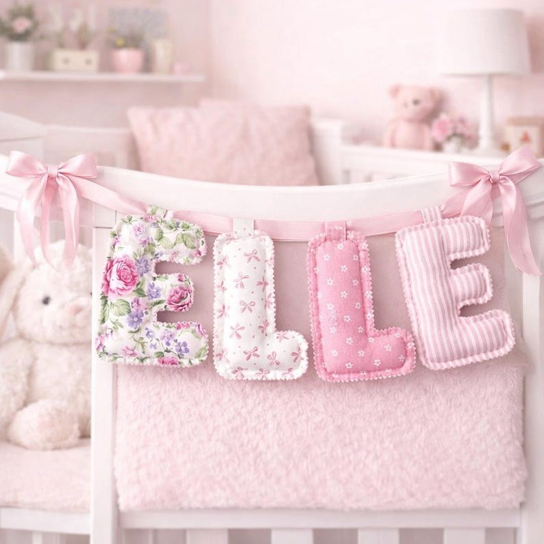 May include: A decorative fabric banner spelling the name ELLE, featuring four plush, stuffed letters hanging from a light pink ribbon with bows on each end. The letters display varied patterns including pink floral, white with pink bows, pink with white stars, and pink stripes.
