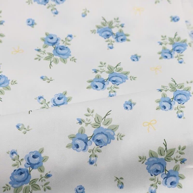 May include: A white fabric featuring a repeating pattern of small blue rose clusters with green leaves and tiny yellow bows scattered throughout the design. The floral print is evenly distributed across the surface of the textile material.