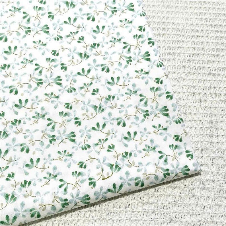 May include: A white fabric featuring a repeating pattern of small, delicate green and light blue floral sprigs. The design consists of tiny leaves and stems scattered across the surface, creating a soft, botanical appearance on the textile.