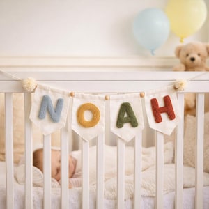 May include: A decorative banner hanging on a white crib, featuring four off-white fabric pennants. Each pennant displays a single, textured, three-dimensional letter in blue, yellow, green, and rust, spelling out the name NOAH with pom-poms.