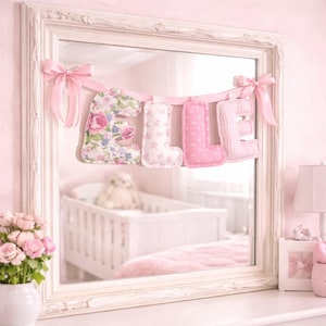 May include: A decorative fabric banner spelling the name ELLE in soft, stuffed letters. Each letter features a different pink pattern, including floral, star, polka dot, and striped designs, suspended by a light pink ribbon with bows.