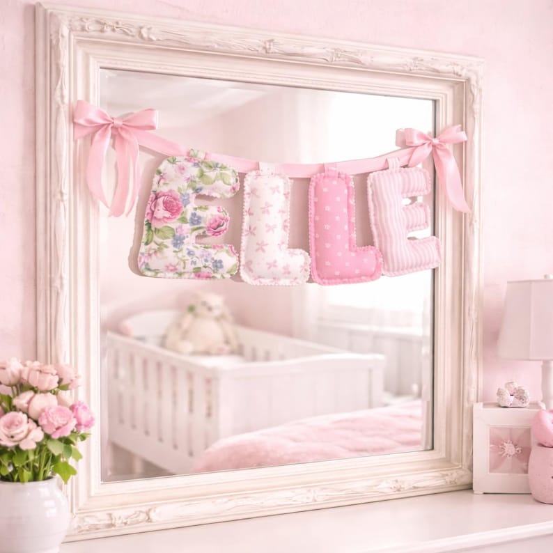 May include: A decorative fabric banner spelling the name ELLE in soft, stuffed letters. Each letter features a different pink pattern, including floral, star, polka dot, and striped designs, suspended by a light pink ribbon with bows.