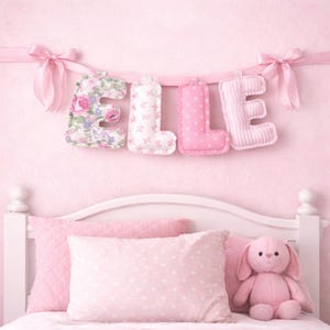 May include: A decorative fabric banner spelling the name ELLE in plush, stuffed letters. Each letter features a different pink pattern, including floral, small bows, polka dots, and stripes, suspended by a pink ribbon with bows at each end.