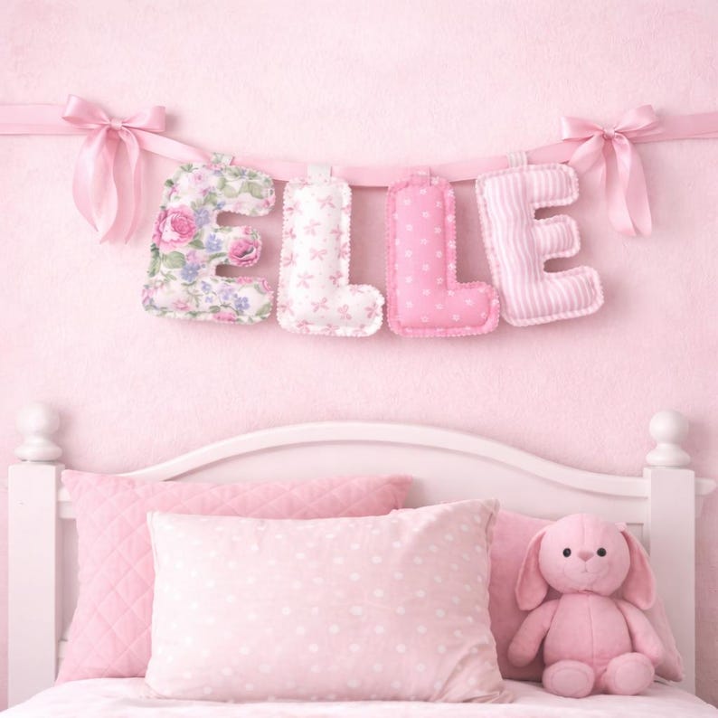 May include: A decorative fabric banner spelling the name ELLE in plush, stuffed letters. Each letter features a different pink pattern, including floral, small bows, polka dots, and stripes, suspended by a pink ribbon with bows at each end.