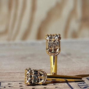 May include: Two gold-colored decorative keys with ornate tops, one standing upright and the other lying down. The keys have a detailed design, possibly nautical-themed, and are set against a light wooden surface.