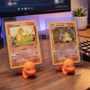 Könnte beinhalten: Zwei Pokémon-Sammelkarten, Charmander und Charizard, in Schutzhüllen. Sie werden von orangefarbenen Charmander-Figuren gehalten. Im Hintergrund befinden sich ein Computermonitor, eine Tastatur und eine kleine Topfpflanze.