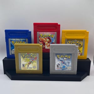 May include: A collection of five vintage Pokémon Game Boy cartridges in blue, red, yellow, gold, and gray. Each cartridge features the Pokémon logo and game-specific artwork. The cartridges are displayed on a black tiered stand.